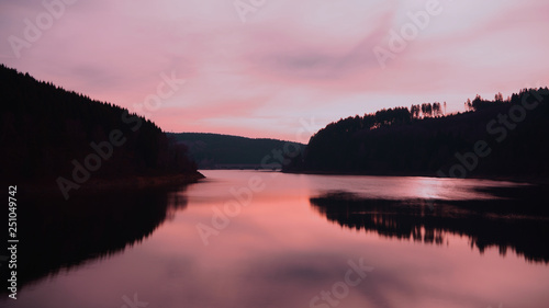 A pink pastel-colored sky and the silhouette of the forest reflect in a calm lake. Oker Dam, Harz mountains, Lower Saxony, Germany.