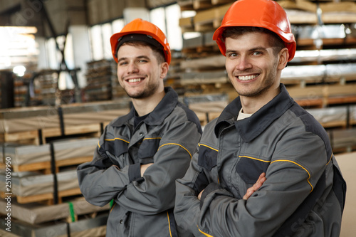 Two young colleagues in gray uniforms and orange helmets standing together with folded arms, smiling, looking at camera and posing on metal stock. Teamwork and collaboration concept.