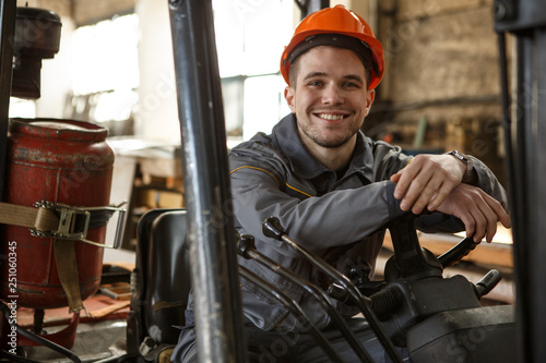 Portrait of metalworker wearing orange hardhat and gray protective suit smiling and looking at camera on stock. Young man sitting on forklift, leaning on steering wheel and enjoying work.