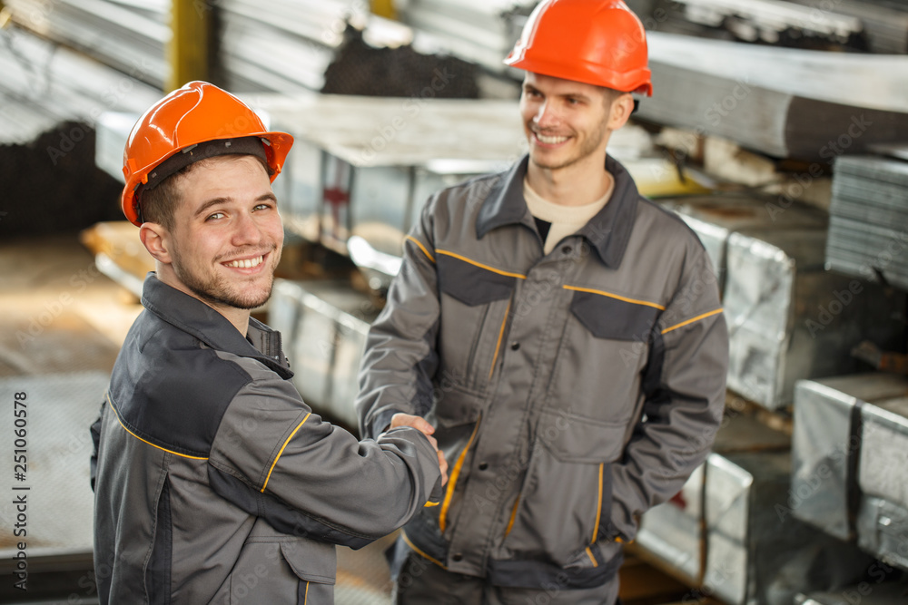 View from above of two cheerful in uniform and helmets shaking hands on metal stock. Happy male worker turning to camera, posing and smiling, his colleague looking at him. Concept of collaboration. View from above of two cheerful in uniform and helmets shaking hands on metal stock. Happy male worker turning to camera, posing and smiling, his colleague looking at him. Concept of collaboration.