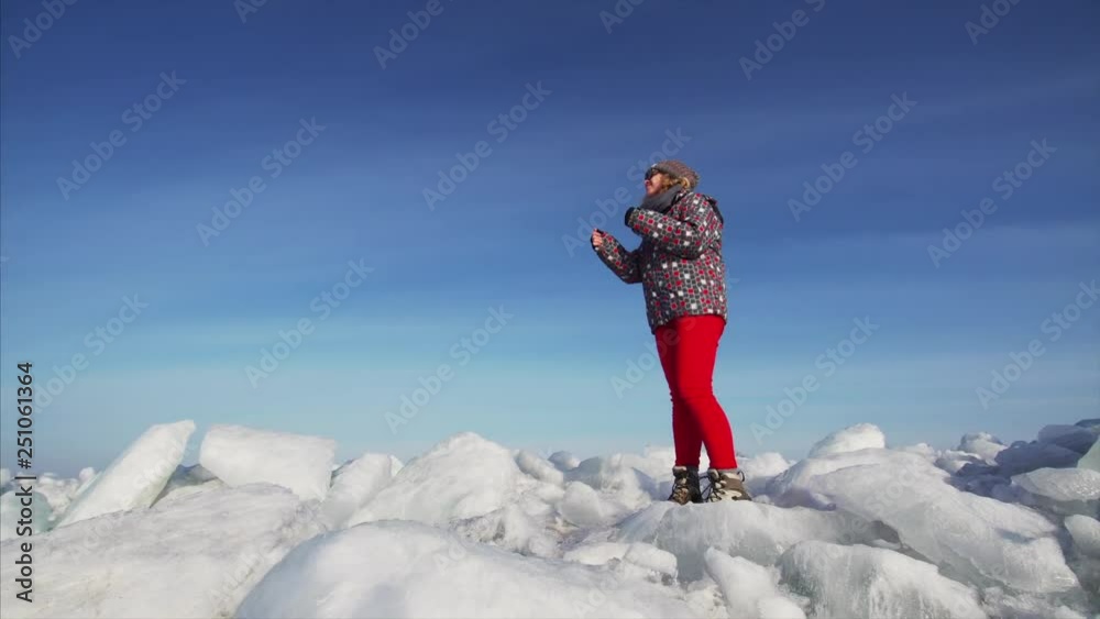 Young woman in glasses is dancing and having fun on big ice blocks. Bright blue sky is on the background. Sunny winter day, lakeside