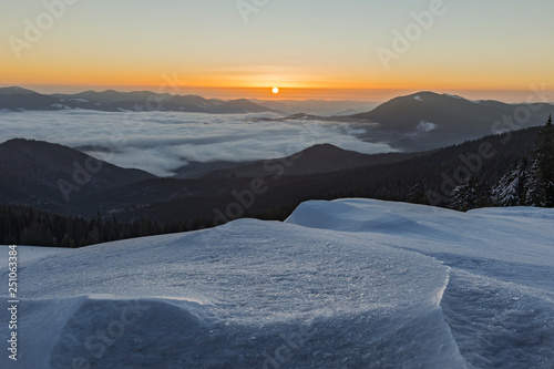 The fantastic scenery of the winter Carpathians with a snow texture in the foreground with dense fog in the mountain valley and the morning sun on the horizon