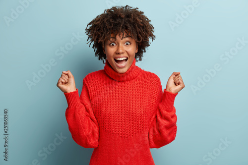 Triumphing overemotive dark skinned woman raises clenched fists, wears red jumper, enjoys success, looks with happiness at camera, recieves promotion, isolated over blue background. Celebration