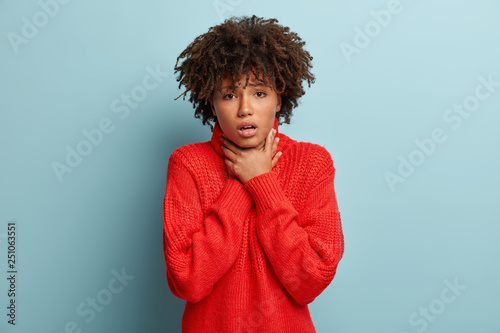 Dissatisfied black woman with curly hair  keeps hands on thore throat  suffers of suffocation  cant breath well  has stressful look  wears oversized red sweater  isolated over blue background.