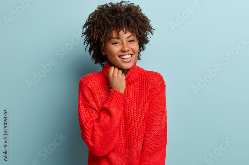 Close up shot of gentle woman with satisfied expression  appealing appearance  keeps hand on collar of red sweater  smiles broadly  has white perfect teeth  isolated over blue background. Emotions