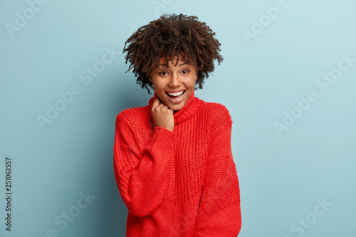 Optimistic glad dark skinned teenage girl enjoys nice friendly conversation, smiles broadly, dressed in warm red sweater, laughs joyfully, isolated over blue background, thinks about perfect vacation