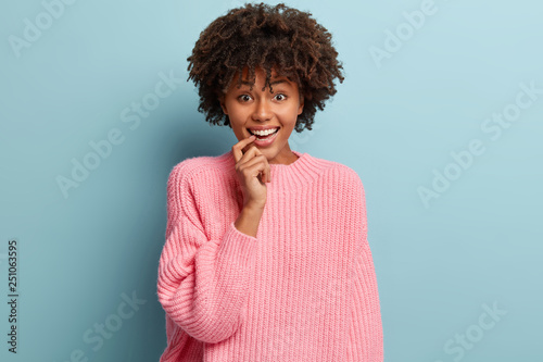 Joyous attractive woman with satisfied expression, keeps fore finger on lips, smiles broadly, wears oversized pink sweater, isolated over blue background, being in high spirit. Happiness concept