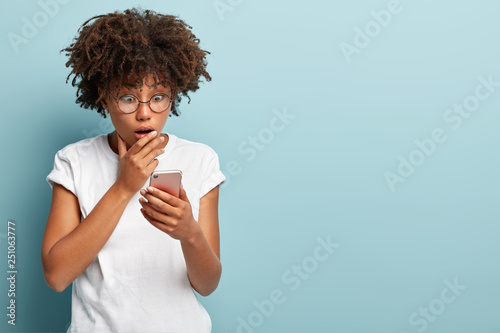 Astonished beautiful woman gazes at modern smartphone, recieves shocking news, covers opened mouth, wears glasses and t shirt, models over blue background with blank space for your promotion photo