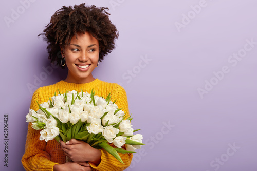 Horizontal shot of good looking black woman with spring flowers bouquet, keeps gaze aside, has curly hairstyle, wears yellow jumper, models over purple background with blank space for information