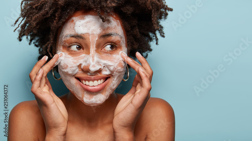 Satisfied beautiful black woman makes massage of face, has white foam, problem of wide pores, cares of skin, keeps gaze aside, smiles positively, models over blue studio wall with free space