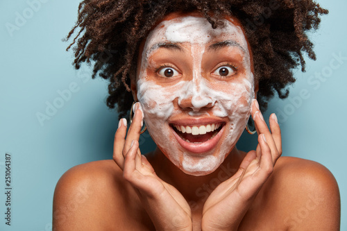 Foto Happy lady wets face with foam, stands in studio with bare shoulders, touches ch