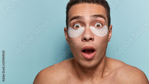 Anti wrinkle treatment concept. Astonished young man has flakes under eyes, white patches, frightened on new therapy, poses topless against blue background, has short hairstyle, being in stupor