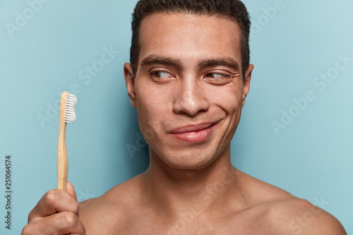 Personal hygiene concept. Pleased Caucasian young man with naked torso, carries bamboo toothbrush, satisfied to lead healthy lifestyle, isolated over blue background. Morning routine. Rear view