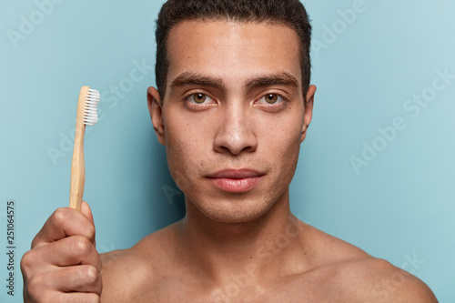 Dental cavity hyginene concept. Serious young Caucasian man with smooth skin, carries wooden toothbrush, has strong muscular nude body, isolated over blue background, ready for brushing teeth photo