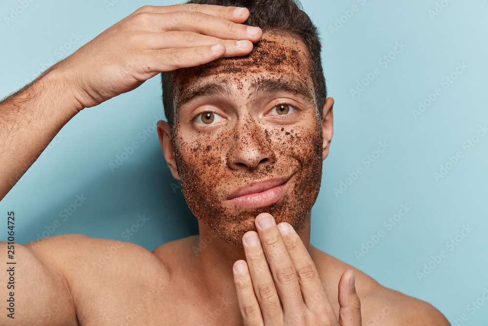 Fototapeta premium Close up photo of displeased young masculinity man has coffee scrub on face, touches chin and forehead, poses topless, models against blue background, visits cosmetologist. Wellness concept.