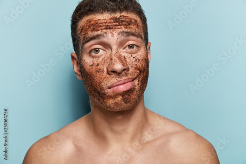 Masculine beauty therapy concept. Young male with coffee scrub mask, purses lips, looks with displeasure, has bare muscular strong body, short dark hairstyle, isolated over blue studio background