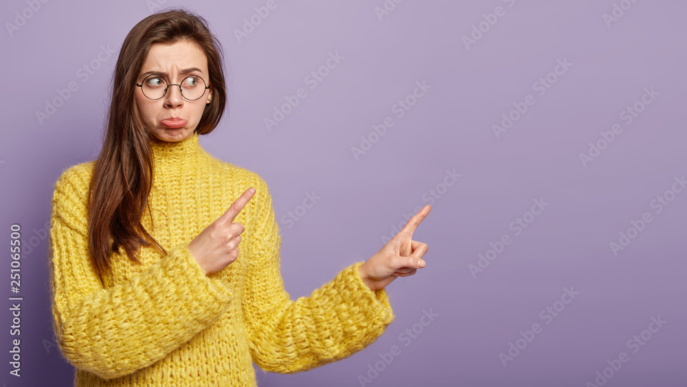 Waist up shot of displeased young woman feels perplexed and frustrated, purses lips, has long hair, points away with both fore fingers, dressed in yellow clothes, isolated on purple background Waist up shot of displeased young woman feels perplexed and frustrated, purses lips, has long hair, points away with both fore fingers, dressed in yellow clothes, isolated on purple background
