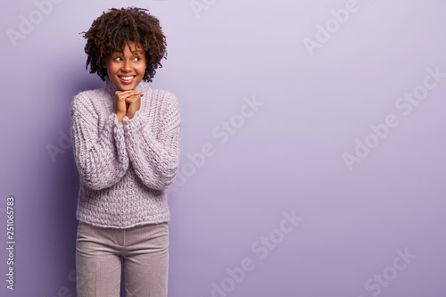 Joyful satisfied woman with curly hairstyle, smiles gently, keeps look away, holds hands together under chin, wears knitted sweater and trousers, isolated over purple background with mockup space