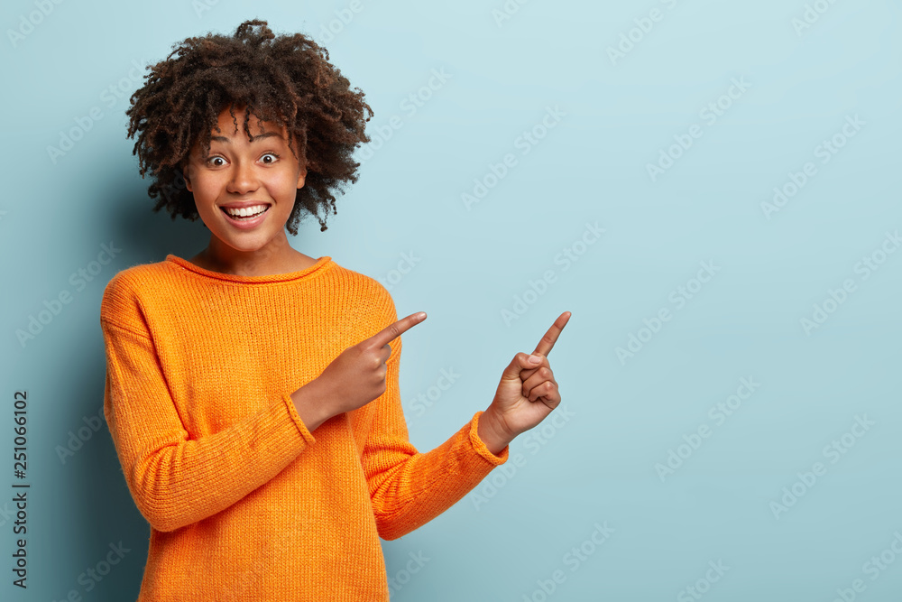 Cheerful lovely young girlfriend with broad smile, shows white teeth, indicates with both fore fingers at blank space, wears orange jumper, isolated over blue wall. Buy this thing on discount or sale - obrazy, fototapety, plakaty 