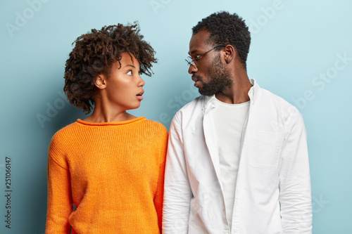 Obraz na plátne Stunned dark skinned female and male partners look surprisingly at each other, forget to accomplish important task, dressed in casual outfit, isolated over blue background