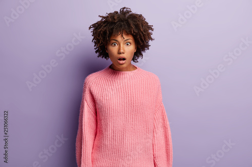Stunned emotional woman being speechless and impressed by shocking news  keeps jaw dropped  dressed in casual oversized pink sweater  isolated over purple background. Scared black girl indoor