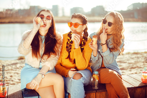 Three girls sit on the beach   near the river  talk  gossip  joke  laugh  smile at womans day. Girls on a hot day eat ice cream. Hipster model wearing sunglasses at summer day. Girl with hairslyle