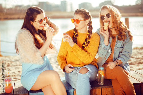 Three girls sit on the beach , near the river, talk, gossip, joke, laugh, smile at womans day. Girls on a hot day eat ice cream. Hipster model wearing sunglasses at summer day. Girl with hairslyle © MoreThanProd