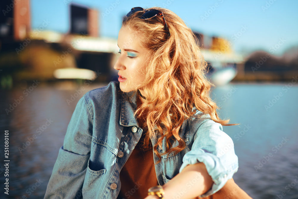 Beautiful girl sitting on the pier near the river. Hipster model in sunglasses with gorgeous  cerly hair. The girl in the denim jacket looks into the distance at sunny day. Summer time. Choker on girl