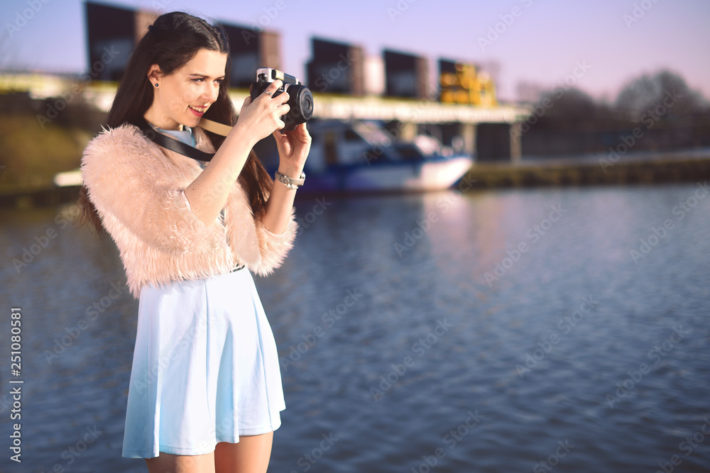 Beautiful girl on the pier near the river. Model in blue dress with long hair. Girl at hot sunny day taking pictures on old camera, Summer vibes. Woman photographer hobby. On the background of yachts