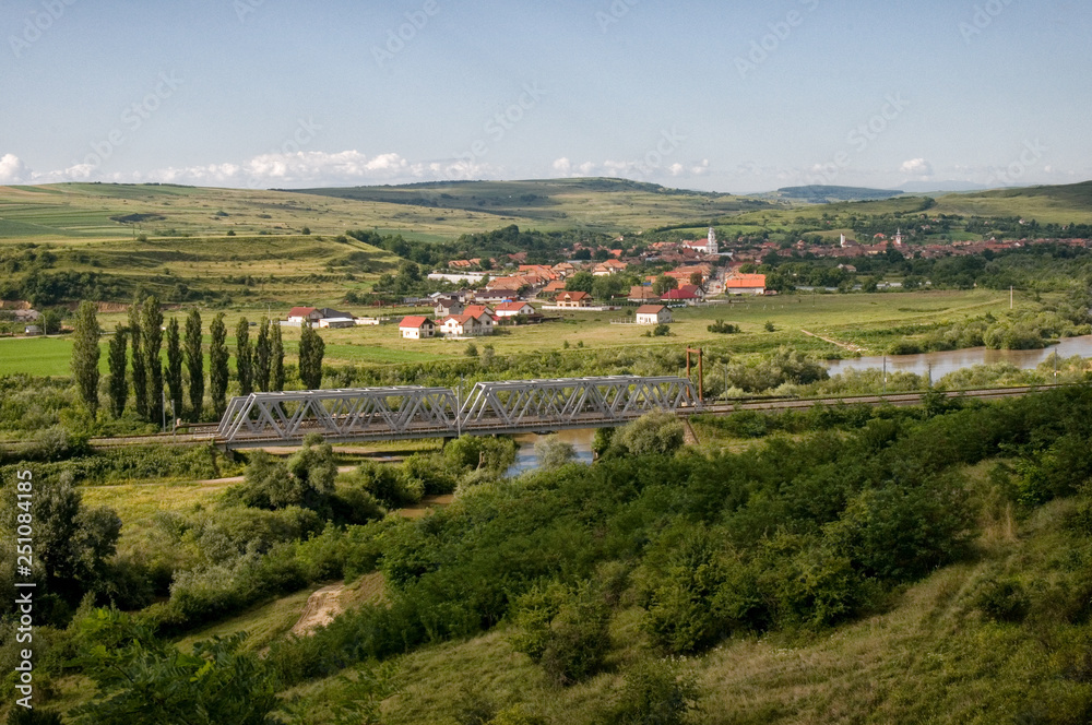A steel train bridge over river in Romania with village in background and vegetation in foreground