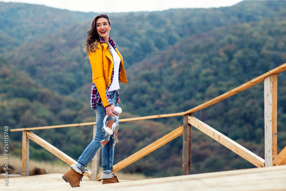 Adorable long hair girl standing at the wooden bridge with forest on the background with headphones and coffer to go on hand Adorable long hair girl standing at the wooden bridge with forest on the background with headphones and coffer to go on hand