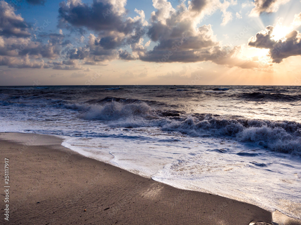 Beach at sunset with choppy sea and waves crashing to shore.