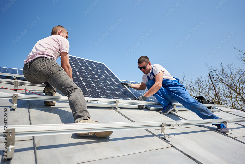 Male workers installing stand-alone solar photovoltaic panel system. Two electricians mounting blue solar module on roof of modern house. Alternative renewable energy concept. Male workers installing stand-alone solar photovoltaic panel system. Two electricians mounting blue solar module on roof of modern house. Alternative renewable energy concept.