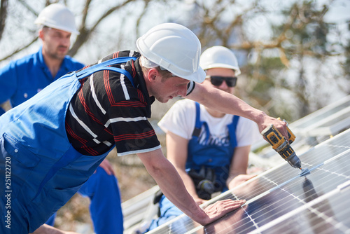 Male team workers installing stand-alone solar photovoltaic panel system using screwdriver. Electricians mounting blue solar module on roof of modern house. Alternative energy ecological concept.