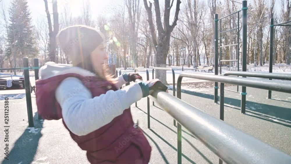 Portrait of girl athlete in a hat with gloves and a warm vest doing exercises on uneven bars at an outdoor sports field in winter on a sunny day Portrait of girl athlete in a hat with gloves and a warm vest doing exercises on uneven bars at an outdoor sports field in winter on a sunny day