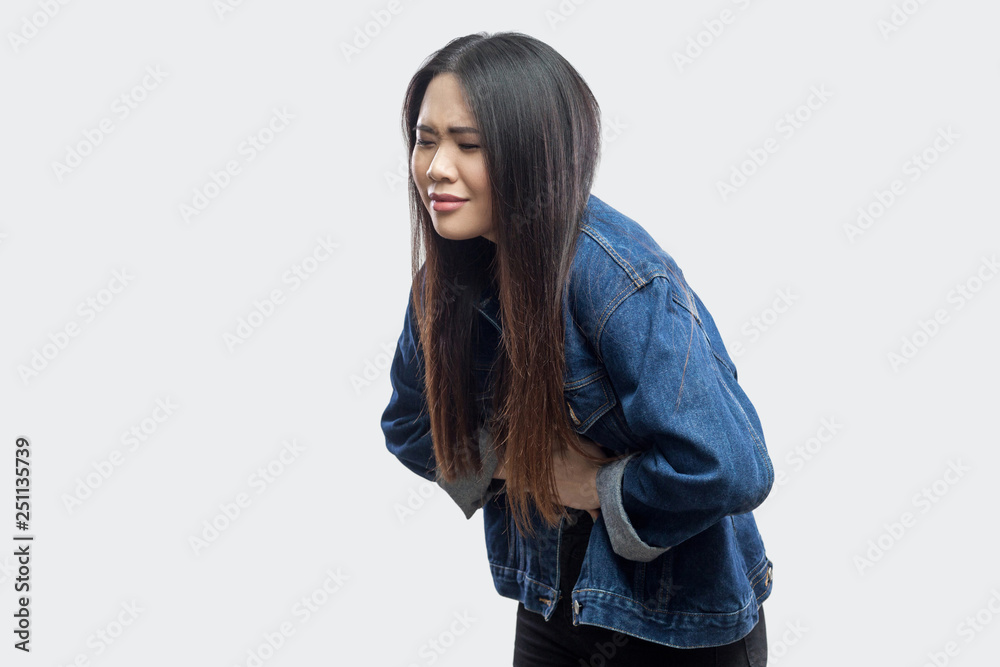 Stomach pain. Side view portrait of beautiful brunette asian young woman in casual blue denim jacket standing and feeling pain on her stomach. indoor studio shot, isolated on light grey background. Stomach pain. Side view portrait of beautiful brunette asian young woman in casual blue denim jacket standing and feeling pain on her stomach. indoor studio shot, isolated on light grey background.
