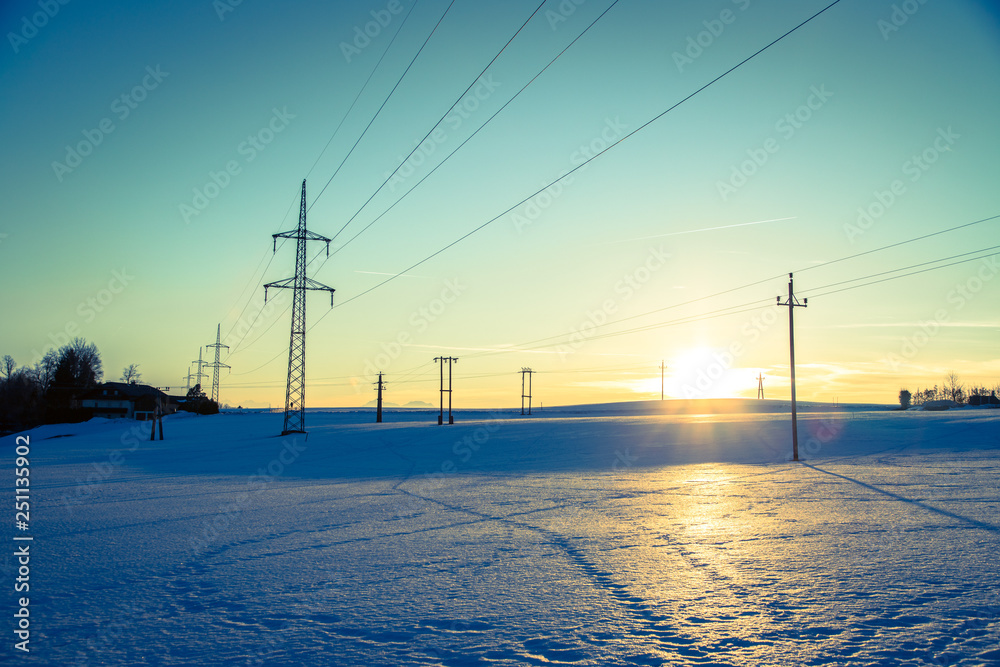 Electrical tower on a snowy field. Sun goes down, orange colors, evening scenery