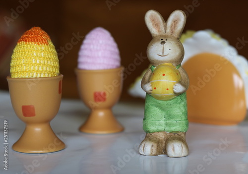 Figurka wielkanocnego zajączka z żółtym jajkiem w łapkach, obok dwie kolorowe pisanki z włóczki w glinianych pojemnikach na jajka, rozmyty pojemnik z serwetkami w tle