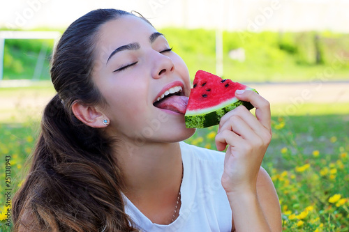 portrait of a girl with dark hair holding a juicy watermelon and licking it, against the background of a green field