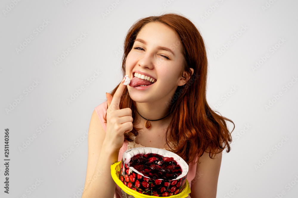 an attractive red-haired young woman holds a cake in her hand and licks a pastry cream off her finger. She have a sweet tooth and love sweets an attractive red-haired young woman holds a cake in her hand and licks a pastry cream off her finger. She have a sweet tooth and love sweets