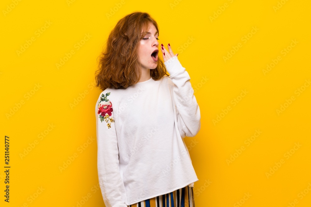 Young redhead woman over yellow wall yawning and covering wide open mouth with hand Young redhead woman over yellow wall yawning and covering wide open mouth with hand