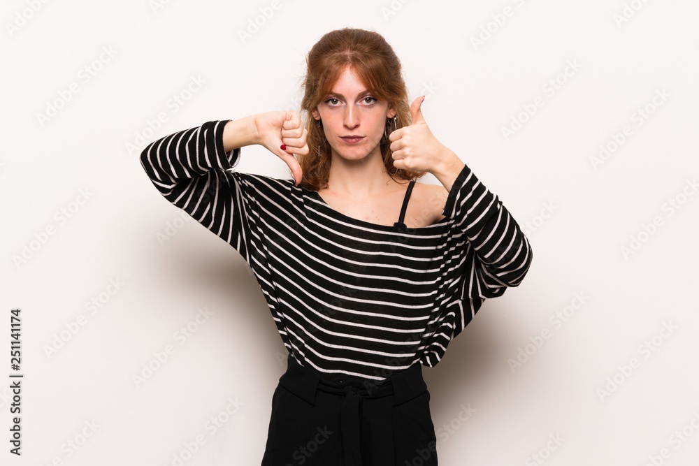 Young redhead woman over white wall making good-bad sign. Undecided between yes or not