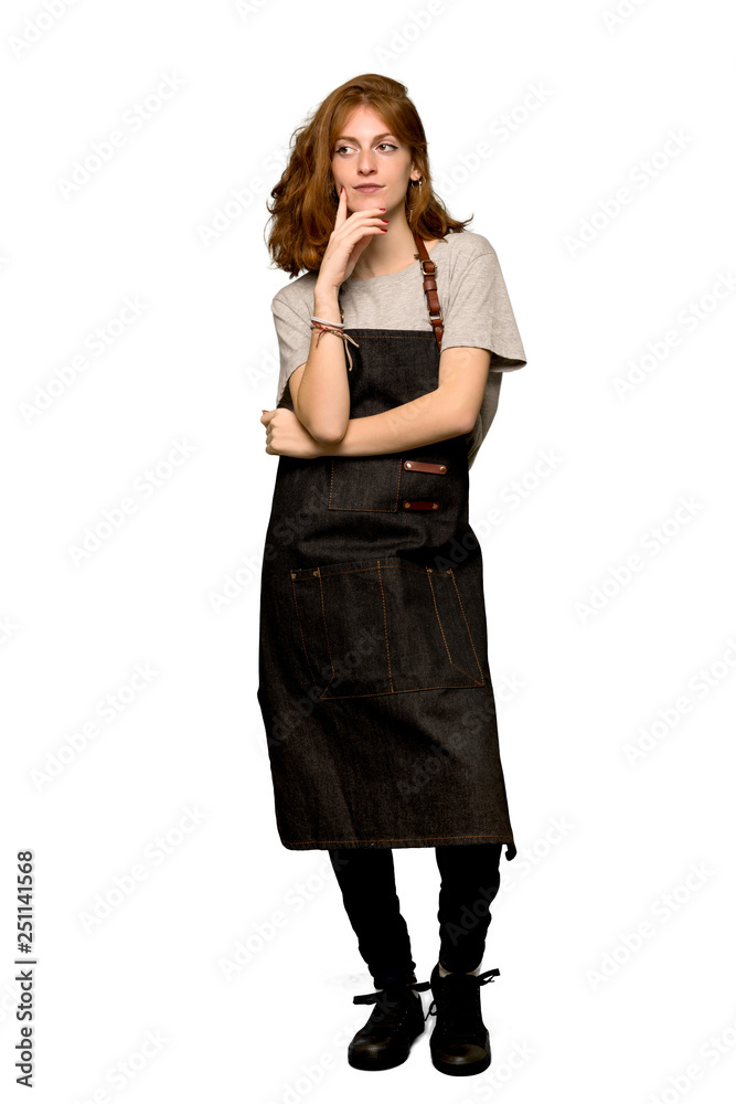 A full-length shot of a Young redhead woman with apron thinking an idea while looking up over isolated white background A full-length shot of a Young redhead woman with apron thinking an idea while looking up over isolated white background