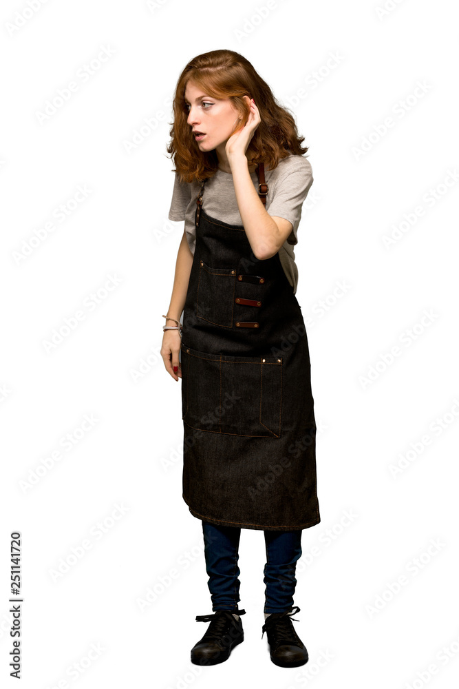 A full-length shot of a Young redhead woman with apron listening to something by putting hand on the ear over isolated white background A full-length shot of a Young redhead woman with apron listening to something by putting hand on the ear over isolated white background