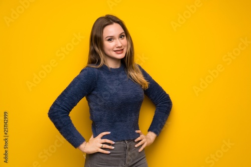 Teenager girl over yellow wall posing with arms at hip and smiling