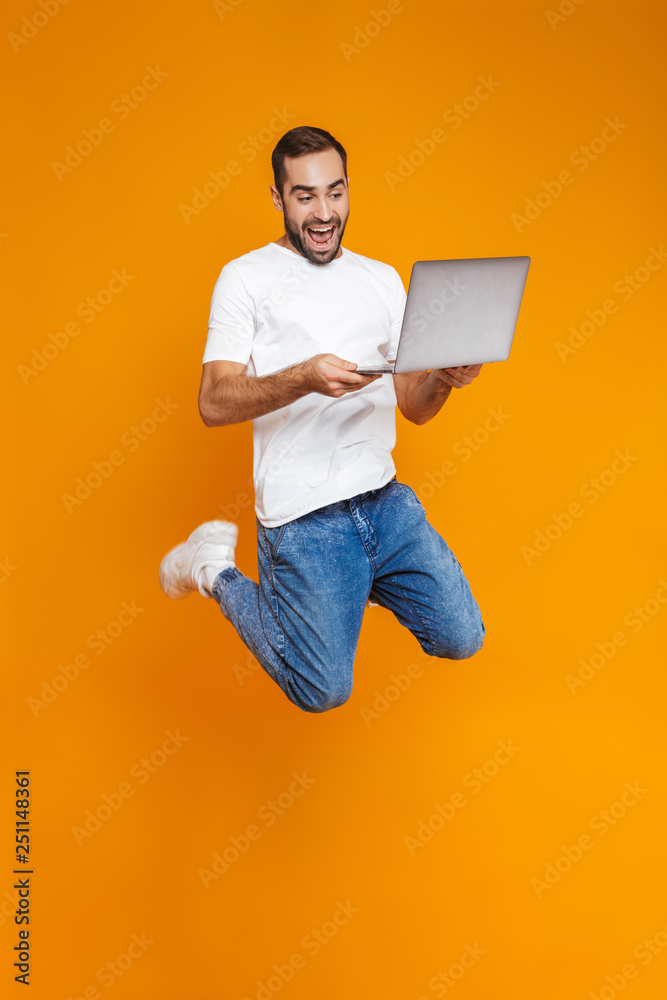 Full length portrait of cheerful man 30s in white t-shirt holding silver laptop, isolated over yellow background