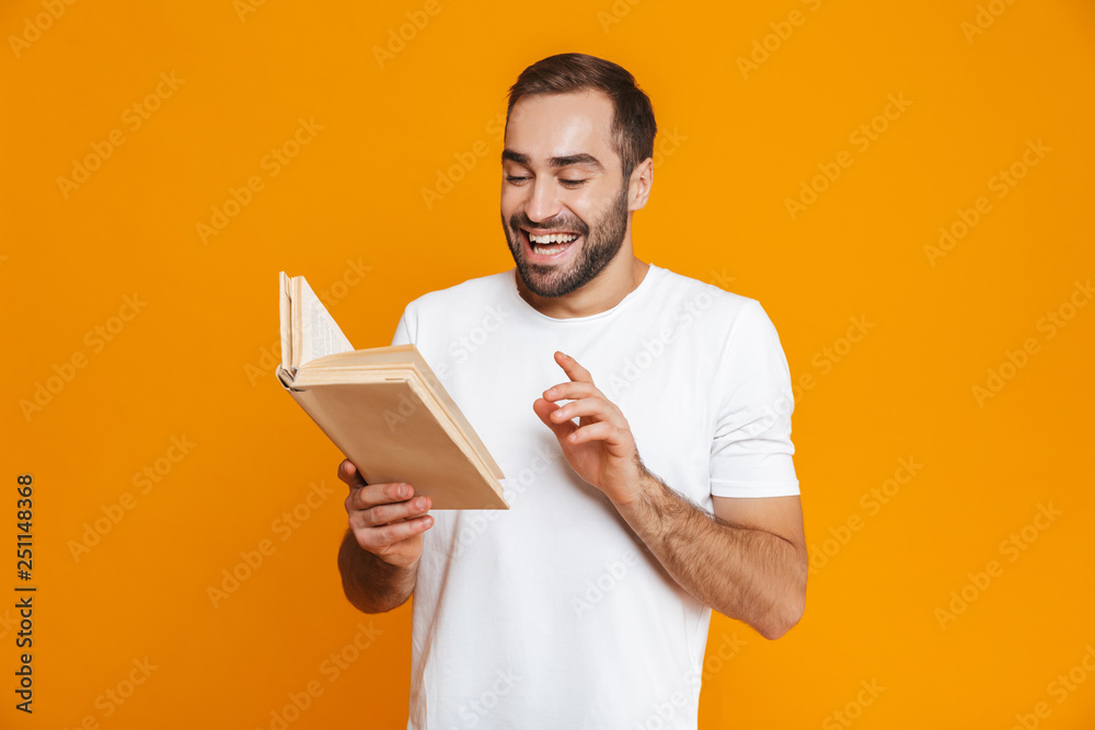 Image of attractive man 30s in white t-shirt holding and reading book, isolated over yellow background Image of attractive man 30s in white t-shirt holding and reading book, isolated over yellow background