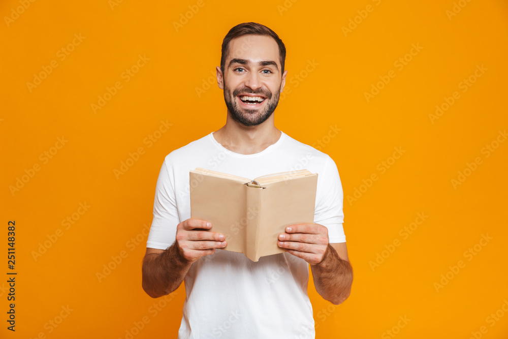 Image of optimistic man 30s in white t-shirt holding and reading book, isolated over yellow background Image of optimistic man 30s in white t-shirt holding and reading book, isolated over yellow background