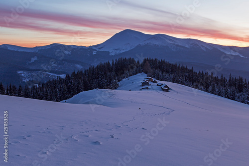Dramatic wintry scene with snowy house. Fantastic colors of the sunset in the winter Carpathians with the shepherds' hut in the foreground and high peaks on the horizon