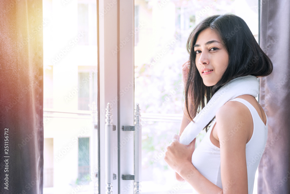 Cheerful smiling young woman 20s black long hair with towel at her neck in the morning standing beside the window while looking to the camera.